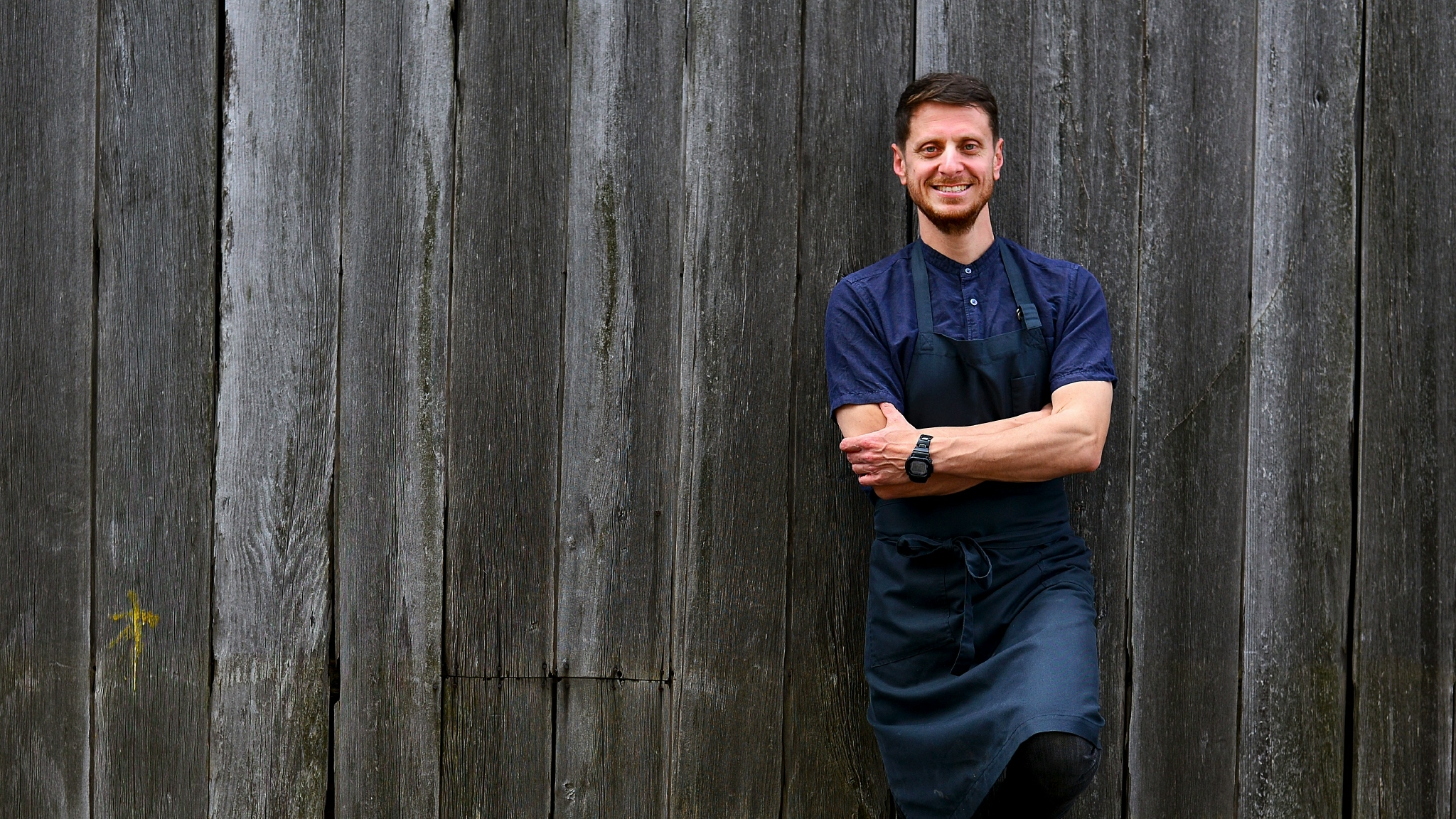 Chef Matteo Alberti stands arms crossed against a a wooden wall